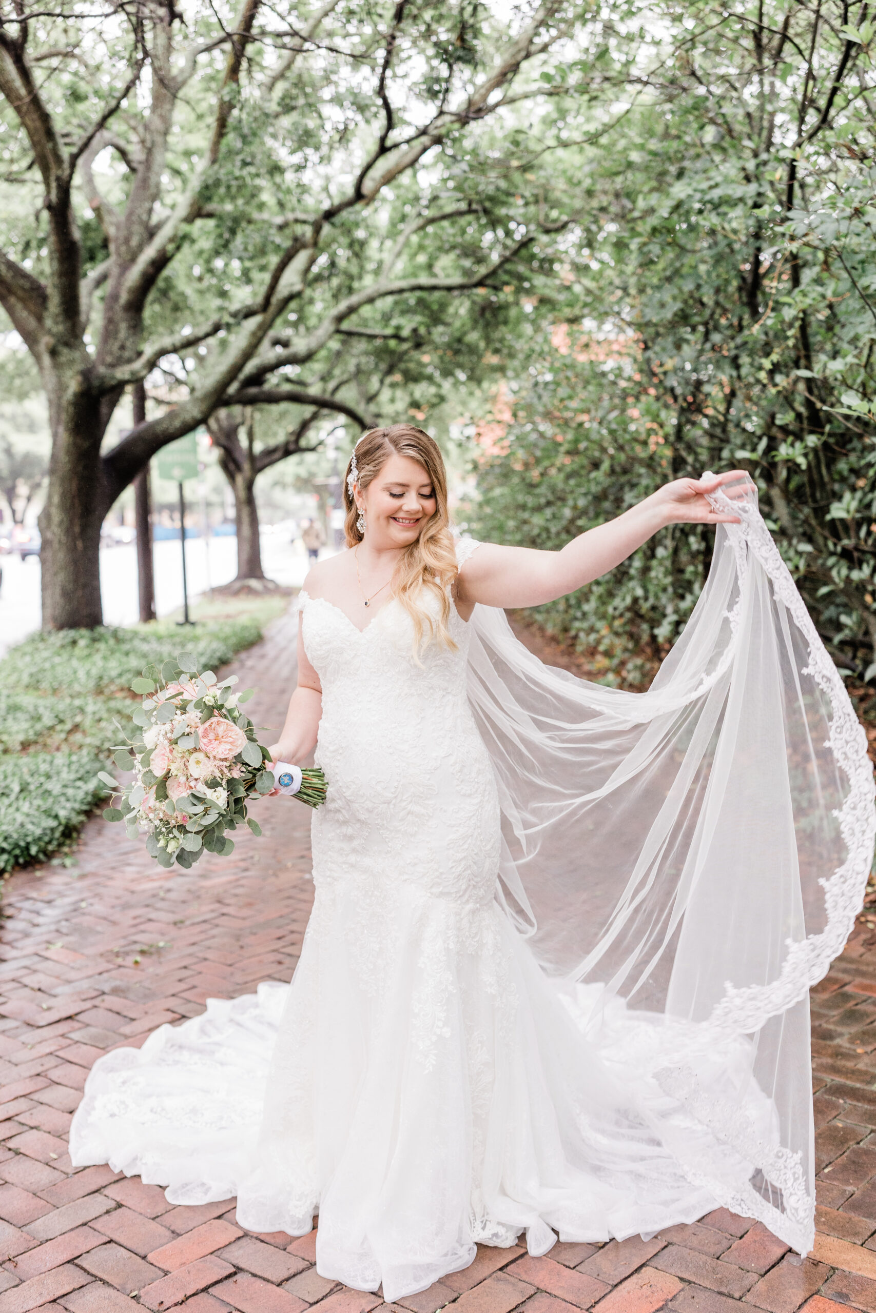 A bride is holding her veil in front of a brick walkway.